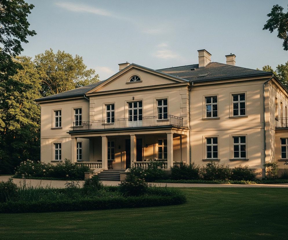 Duży, beżowy budynek w klasycznym stylu z ciemnym dachem i symetrycznymi oknami w białych ramach dominuje centralną część kadru. Przed frontowym wejściem znajdują się schody prowadzące do portyku z kolumnami, nad którym rozciąga się balkon z czarną, metalową balustradą. Po obu stronach budynku rosną bujne drzewa o ciemnozielonych liściach, a przed nim rozpościera się jasnozielony trawnik, z przylegającą do niego żwirową ścieżką i nisko przyciętymi krzewami. Zachodzące słońce rzuca długie cienie na elewację budynku i oświetla jego prawą część ciepłym, złotym światłem.