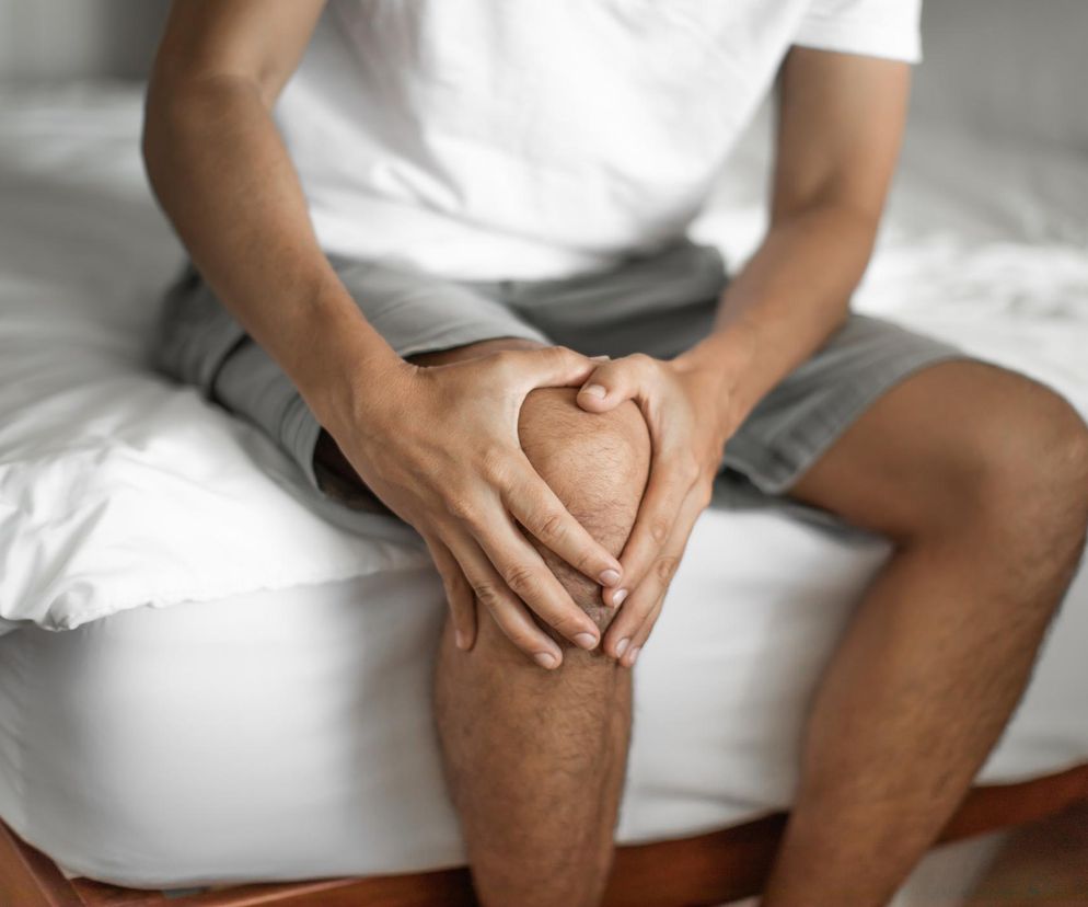 Mężczyzna w jasnej koszulce i szarych spodenkach siedzi na łóżku, trzymając dłonie na bolącym kolanie, co symbolizuje poranny ból i sztywność stawów, jeden z objawów reumatoidalnego zapalenia stawów (RZS). Więcej o chorobach stawów przeczytasz na Poradnik Zdrowie.