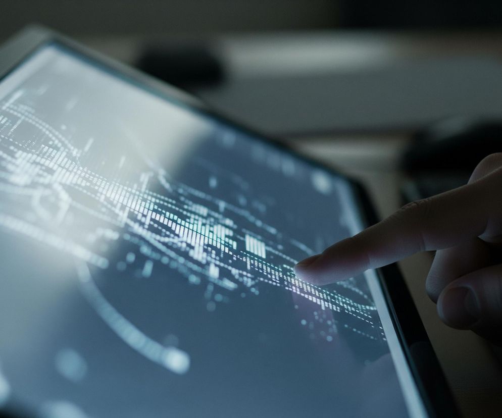 Ekran dotykowy, prawdopodobnie tabletu lub laptopa, wyświetla abstrakcyjny, biało-niebieski wykres danych, z dużą ilością małych kropek i słupków. Prawa dłoń osoby wskazuje palcem wskazującym na centralny punkt na ekranie. Tło jest rozmazane i ciemne, widać na nim fragment klawiatury i inne elementy biurka.