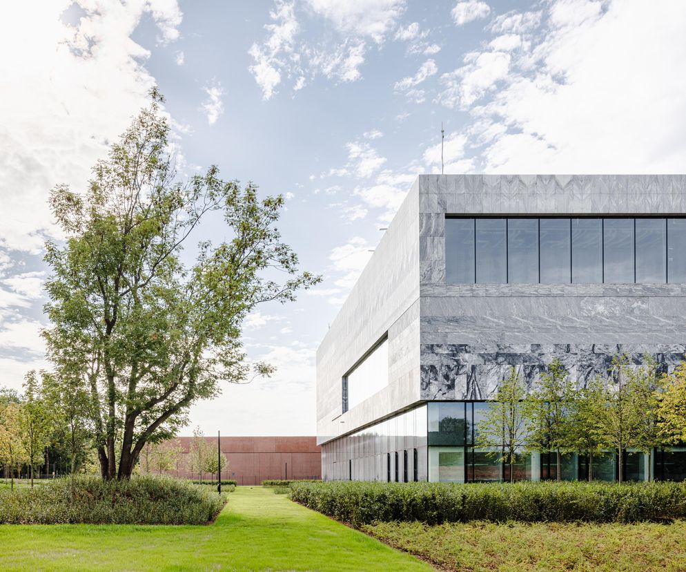 Nowoczesny budynek z szarego kamienia z dużymi oknami, otoczony zielonym trawnikiem i drzewami, w tle widać ceglastą ścianę. To fragment Cytadeli Muzeów autorstwa WXCA, opisywanej na portalu Architektura Murator Plus.
