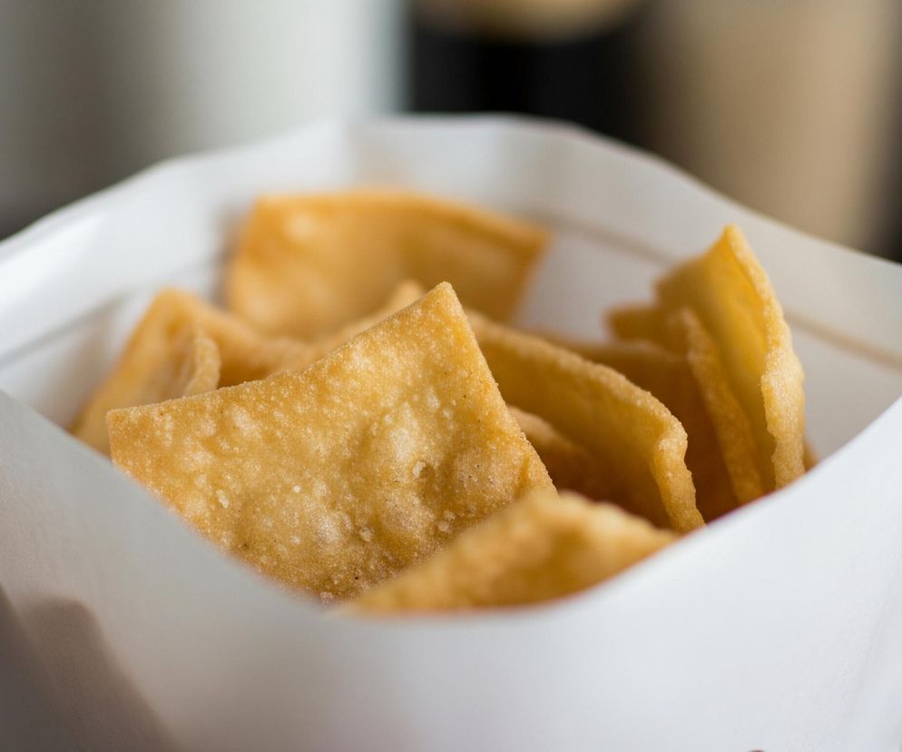 Białe papierowe opakowanie wypełnione jest wieloma złocistymi, chrupiącymi kawałkami smażonego ciasta, które mają nieregularne kształty i widoczne pęcherzyki powietrza na powierzchni. Kawałki ciasta są ułożone warstwowo, a te z przodu są wyraźniejsze niż te w głębi. Tło jest rozmyte, z widocznymi odcieniami szarości, brązu i czerni.