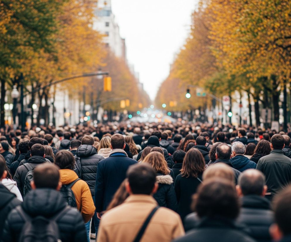 Duży tłum ludzi idzie szeroką aleją wysadzaną drzewami o żółto-zielonych liściach. Postaci, głównie ujęte od tyłu, wypełniają całą szerokość ulicy, z ostrością na tych najbliżej widza. W tle widać niewyraźne budynki i sygnalizację świetlną z zielonymi światłami.