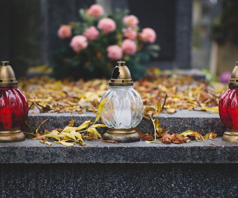 Trzy znicze stojące na nagrobku, symbolizujące pamięć o zmarłych podczas Wszystkich Świętych. Dwa czerwone i jeden przezroczysty lampion oświetlają jesienne liście i kwiaty w tle. Dowiedz się więcej o grobing i jego kosztach na portalu Super Biznes.