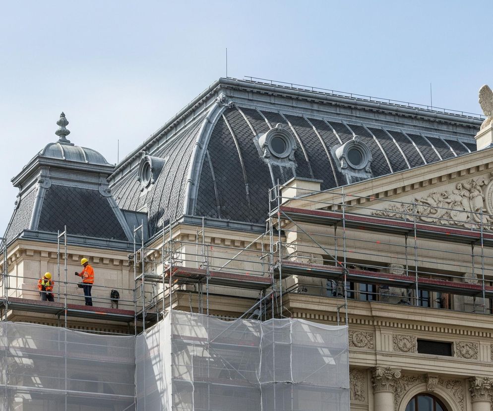 Budynek z mansardowym dachem w kolorze ciemnoszarym, pokrytym drobnymi, romboidalnymi dachówkami, jest widoczny na tle błękitnego nieba. Na dachu znajdują się ozdobne lukarny o okrągłych oknach, a także wieżyczki z szarymi, metalowymi zdobieniami. Fasada budynku, utrzymana w jasnym, kremowym odcieniu, jest bogato zdobiona rzeźbionymi detalami, gzymsami i kolumnami. Przed częścią budynku stoi rozstawione rusztowanie, częściowo zakryte białymi, półprzezroczystymi siatkami. Na jednym z poziomów rusztowania widoczni są dwaj pracownicy w pomarańczowych kurtkach i żółtych kaskach, a obok nich mały, ciemny pies. Na szczycie prawej części budynku znajduje się kamienna rzeźba uskrzydlonej postaci.
