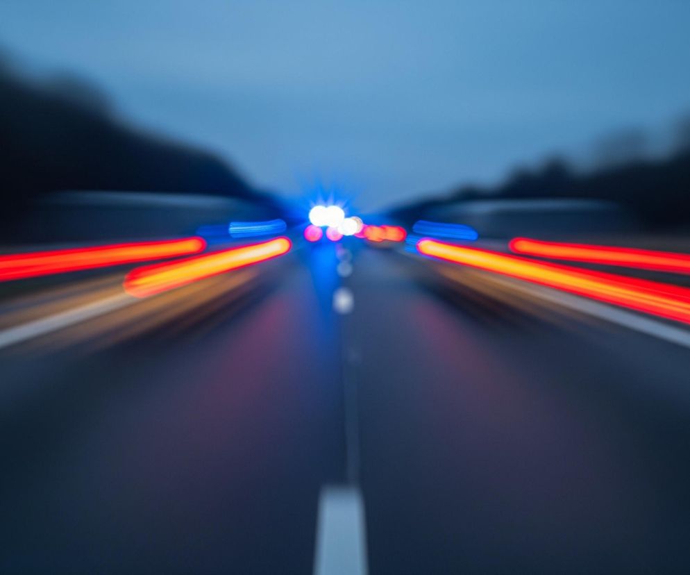 Głównym obiektem na zdjęciu jest droga biegnąca w perspektywie, rozmyta w ruchu. Na ciemnym asfalcie widoczne są intensywne smugi światła: dwie czerwone, ciągnące się po obu stronach drogi, oraz dwie krótsze, pomarańczowo-żółte, położone bliżej środka jezdni. Centralnie, w oddali, znajdują się jasne, rozproszone światła w kolorze niebieskim i białym. Droga jest zaznaczona białą, przerywaną linią pośrodku oraz ciągłymi liniami na krawędziach. W tle, po obu stronach drogi, rozciągają się ciemne, rozmyte kontury drzew lub zalesionych obszarów, a nad nimi widać niebieskawo-szare niebo.