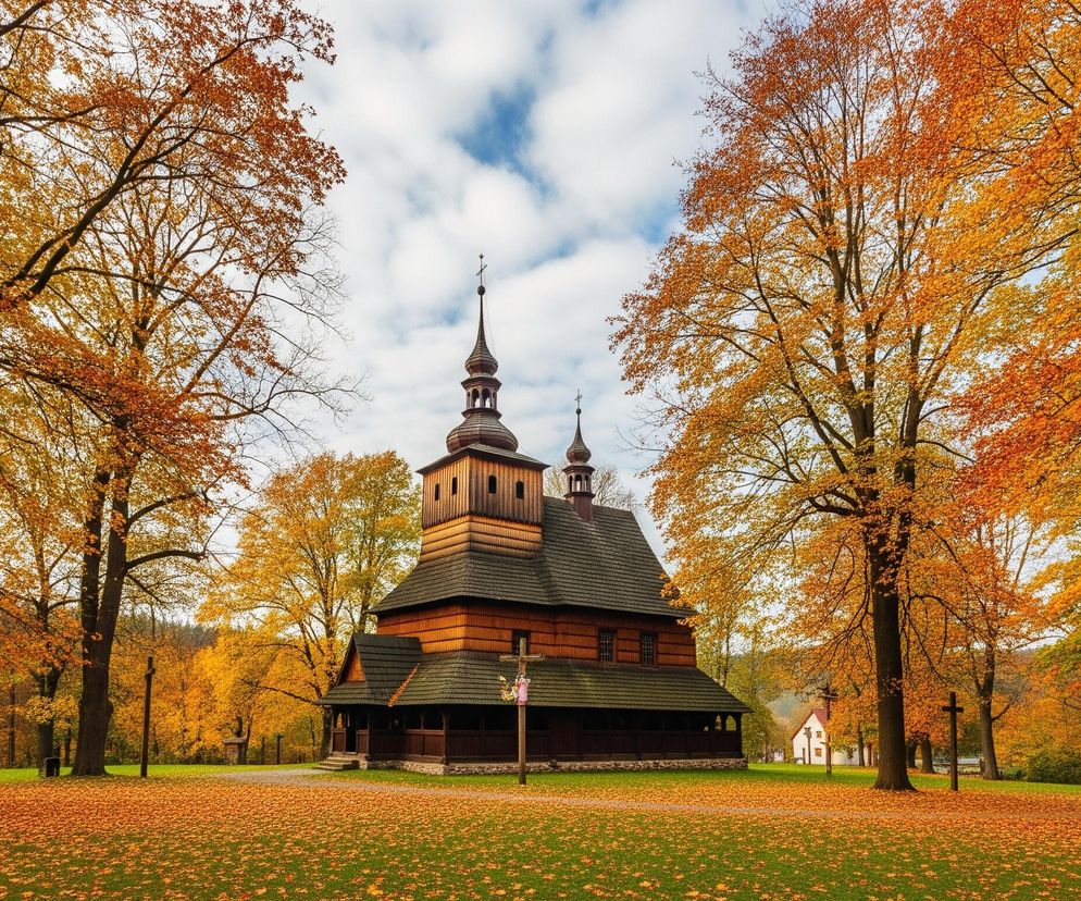 Centralnie, pośrodku obrazu, stoi drewniany kościół o ciemnych, szarych dachach i brązowych, częściowo jaśniejszych ścianach. Kościół posiada dwie wieże, z których większa zwieńczona jest spiczastą iglicą z krzyżem. Obiekt otoczony jest gęstym lasem drzew liściastych o intensywnie pomarańczowo-żółtych liściach, charakterystycznych dla jesieni. Zielona trawa na pierwszym planie pokryta jest obficie opadłymi, pomarańczowymi liśćmi, tworzącymi jednolitą warstwę. Niebo jest częściowo zachmurzone, z wyraźnymi białymi chmurami na niebieskim tle.