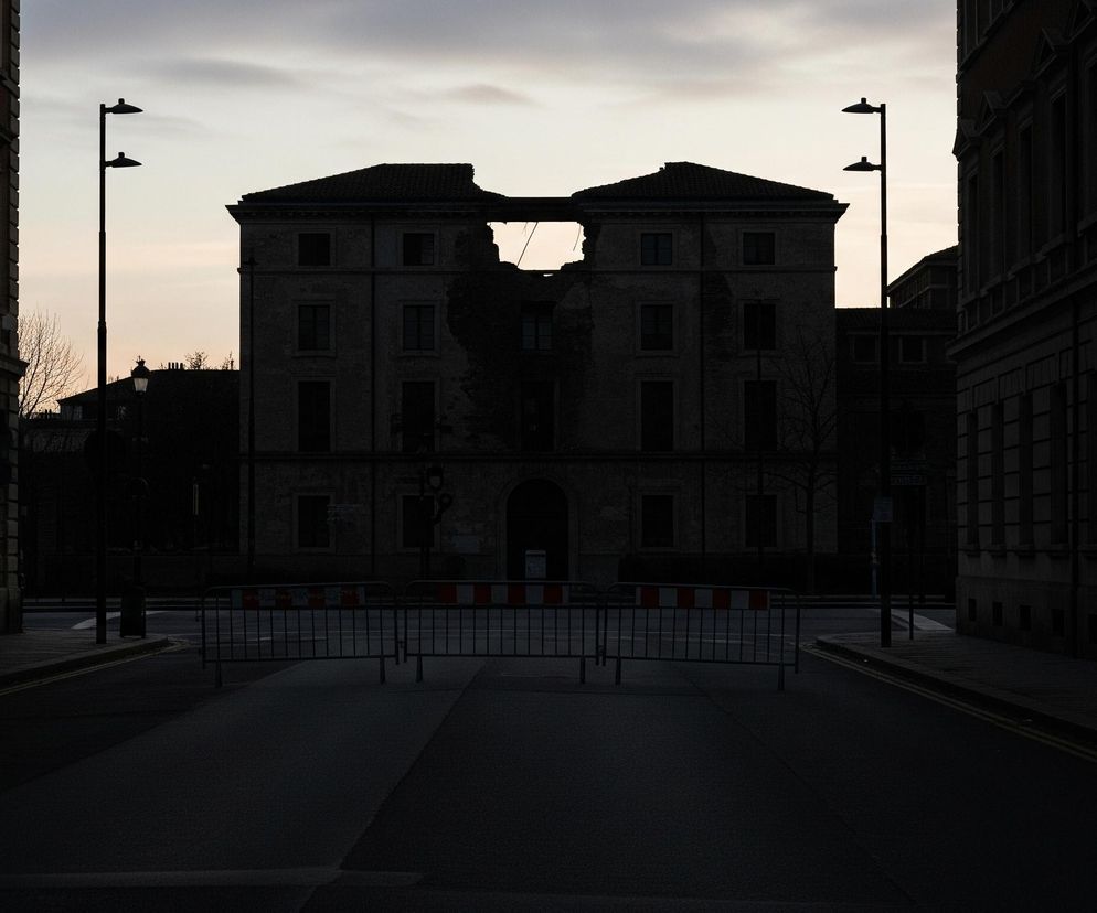 Centralnie, pod wieczornym niebem, widnieje częściowo zrujnowany budynek z pustą, wyrwaną przestrzenią w dachu i górnej części fasady, gdzie widać wnętrze konstrukcji. Obiekt otoczony jest po bokach przez dwa nieuszkodzone budynki o podobnej architekturze, z wyraźnymi oknami i elementami dekoracyjnymi. Na pierwszym planie, na ulicy, znajduje się rząd metalowych barierek z biało-pomarańczowymi elementami, oddzielających obserwatora od głównego obiektu. Wszystkie budynki są ciemne, niemal czarne w cieniu, kontrastując z jaśniejszym, bladoniebieskim i szarym, zachmurzonym niebem, które rozświetla górną część sceny.