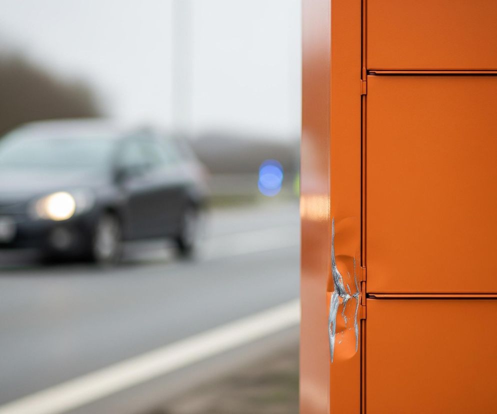 Po prawej stronie kadru widoczny jest pionowy, pomarańczowy słupek lub skrzynka, posiadający wgniecenie i zdarte lakierowanie, ukazujące srebrny metal. Na jego powierzchni znajdują się poziome linie dzielące go na segmenty, a także element przypominający wyświetlacz z częściowo widocznym tekstem i schematycznym rysunkiem w prawym górnym rogu. W tle, po lewej stronie, rozmyty jest ciemnoszary samochód z zapalonymi jasnymi światłami mijania, poruszający się po drodze z białymi pasami. Dalej, za samochodem, widać niewyraźne, niebieskie światło oraz zamazane drzewa lub krzewy.