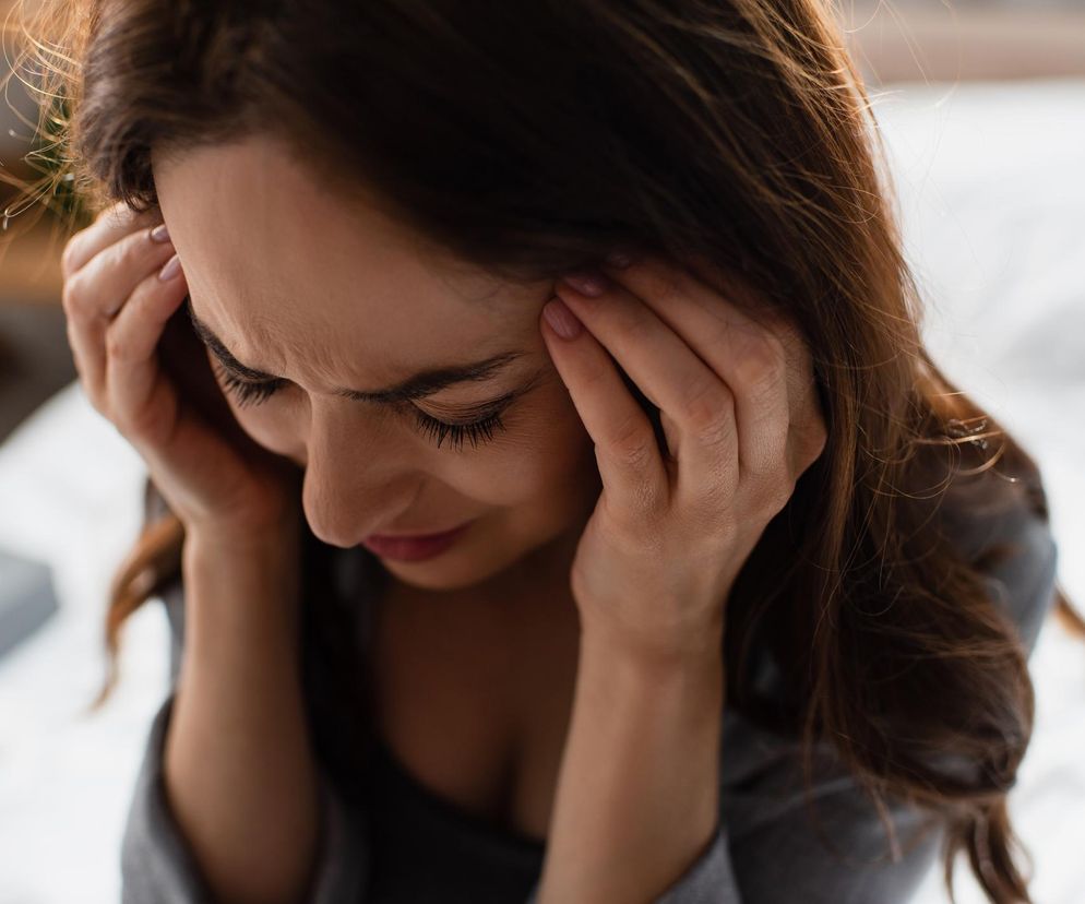 Kobieta z brązowymi włosami cierpiąca na silny ból głowy, masująca skronie. Jej zaciśnięte powieki i ściągnięte brwi wskazują na duży dyskomfort. To zdjęcie ilustruje negatywny wpływ BPA na zdrowie, o którym możesz przeczytać na portalu Poradnik Zdrowie.