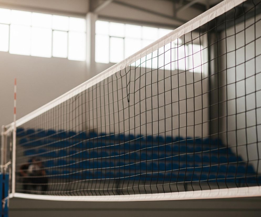 Na pierwszym planie, centralnie, rozciągnięta jest czarna siatka sportowa z białymi obszyciami u góry i na dole. Za siatką widoczne są puste niebieskie trybuny sali gimnastycznej, a w tle duże prostokątne okna, przez które wpada jasne, rozproszone światło. Po lewej stronie siatki widać fragment słupka z biało-czerwonym znacznikiem, a w prawym dolnym rogu obrazu zamazana sylwetka osoby.