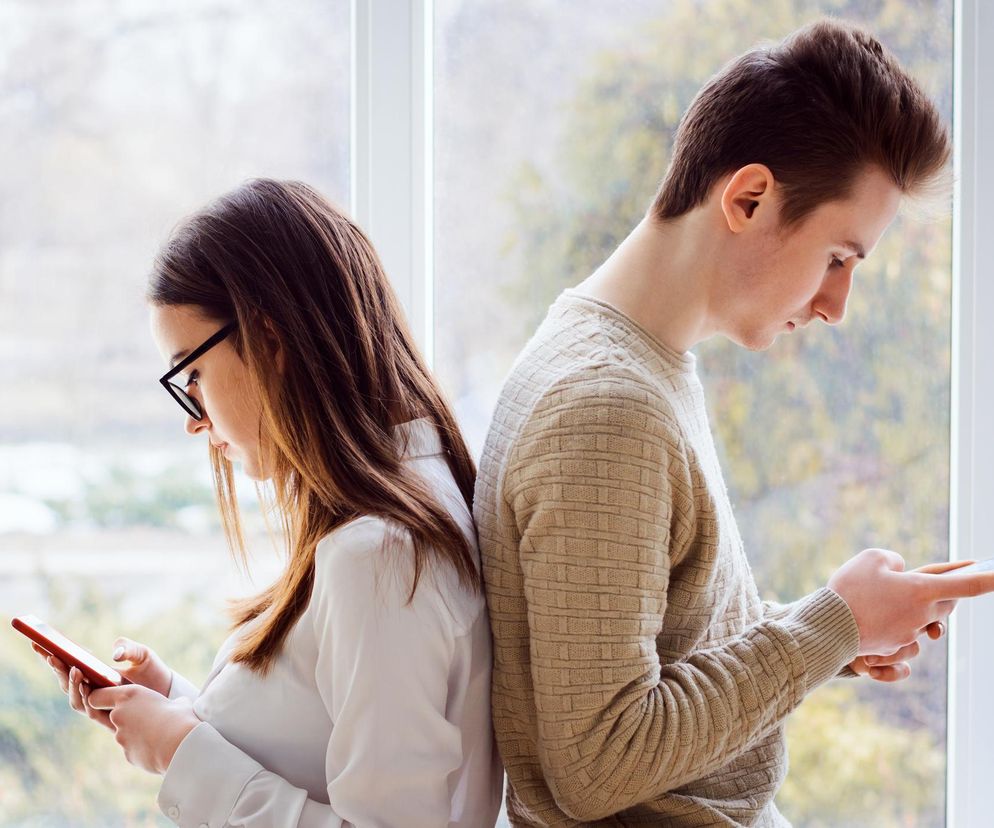 Młoda kobieta w okularach i mężczyzna, oboje z głowami pochylonymi nad smartfonami, stoją do siebie tyłem, opierając się plecami o parapet, z widokiem na niewyraźny krajobraz za oknem. To ilustracja problemu postawy FHP, która może prowadzić do bólu karku i problemów z oddychaniem. Więcej o tym przeczytasz na Poradnik Zdrowie.