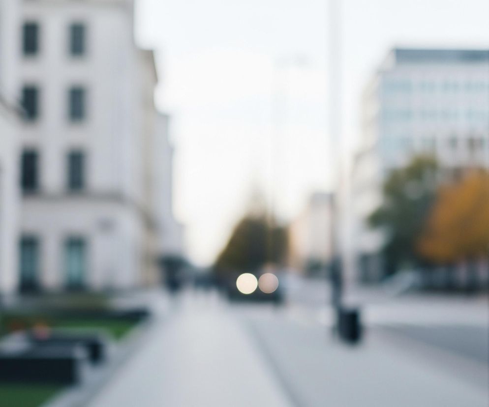 Ulica miasta jest rozmyta, dominują jasne, stonowane barwy. Po lewej stronie widoczne są fragmenty wysokich budynków w odcieniach bieli i szarości, z ciemnymi prostokątnymi oknami. Po prawej stronie widać czarną, pionową latarnię uliczną oraz rozmyte kontury innych budynków i drzew z pomarańczowo-brązowymi liśćmi. W centrum obrazu znajduje się chodnik prowadzący w głąb ulicy, a w oddali majaczą rozmyte światła samochodów.