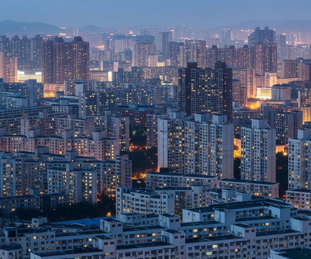 Rozległy krajobraz miejski wypełniają liczne budynki mieszkalne o różnych wysokościach, dominują wieżowce w odcieniach szarości, bieli i beżu. Wiele okien budynków świeci ciepłym, żółtopomarańczowym światłem, tworząc plamy jasności na tle ciemniejącego nieba. W tle, na horyzoncie, widać zarysy gór w niebieskawych odcieniach, a pomiędzy nimi rozpościera się mglista łuna świateł odległych zabudowań.