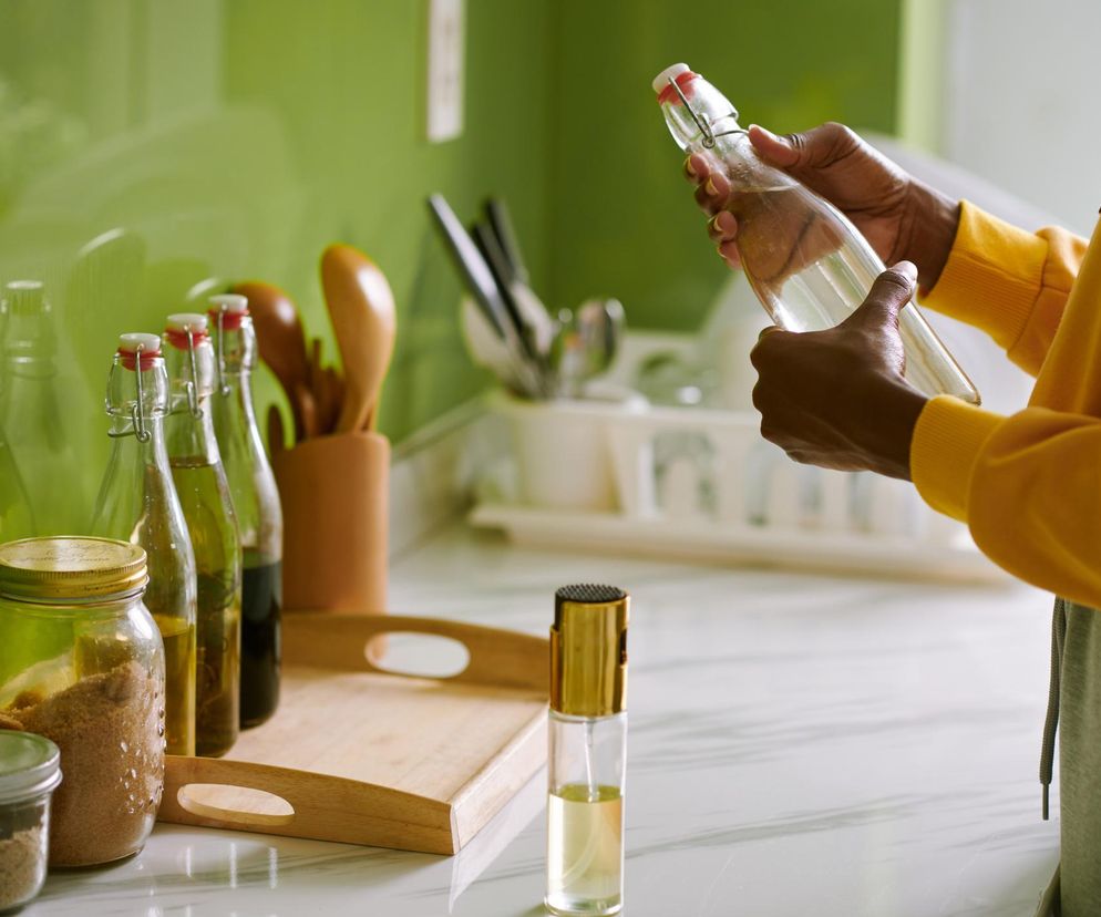 Osoba o ciemnej karnacji w żółtej bluzie, trzymająca szklaną butelkę z octem jabłkowym. Na blacie widoczne są inne butelki i słoiki. Zdjęcie symbolizuje fałszywe obietnice o odchudzaniu octem jabłkowym, o czym można przeczytać na portalu Poradnik Zdrowie.