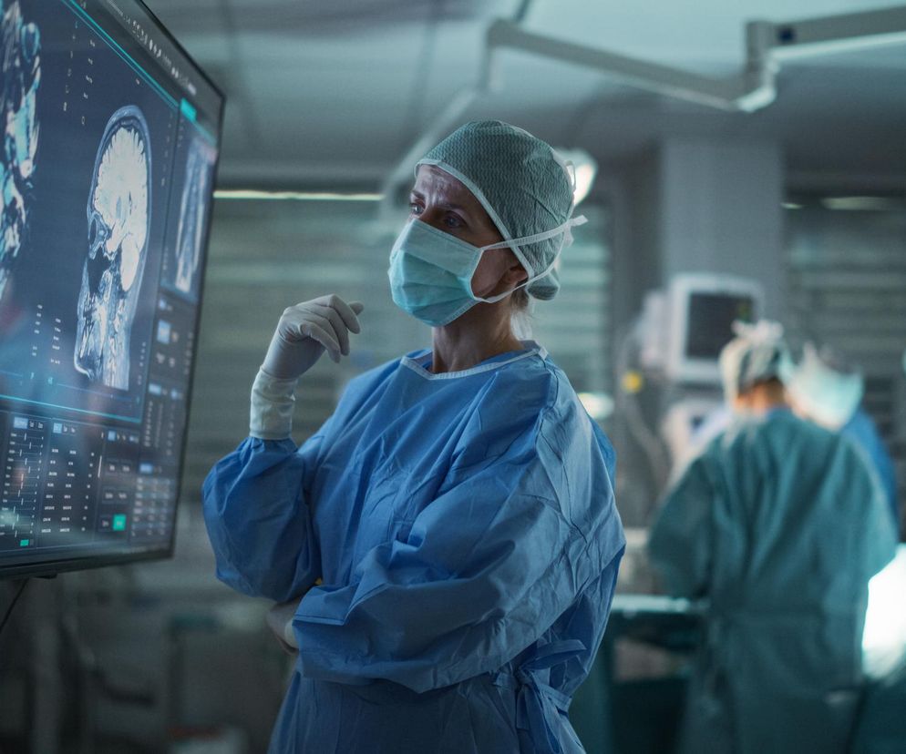 Lekarka w masce chirurgicznej, czepku i fartuchu, z uwagą oglądająca na monitorze wyniki rezonansu magnetycznego mózgu, obok niej niewyraźne sylwetki innego personelu medycznego. Zdjęcie ilustruje problem błędów medycznych i potrzebę elektronicznego rejestru zdarzeń niepożądanych, o czym przeczytasz na Poradnik Zdrowie.