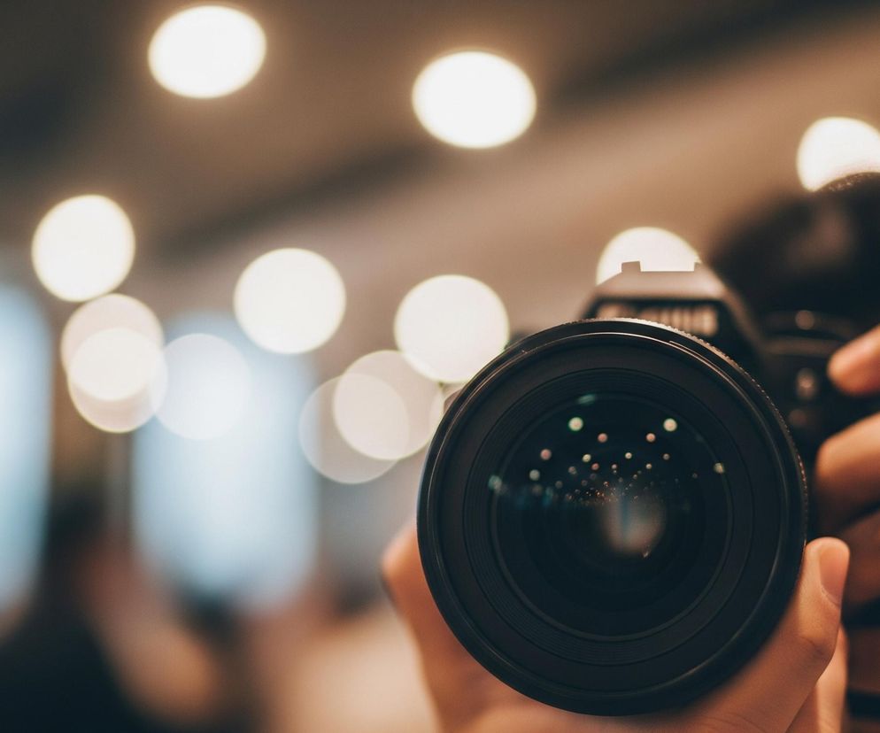 Obiektyw czarnego aparatu fotograficznego dominuje centralną część kadru, trzymany przez dłonie osoby. Na jego powierzchni widoczne są liczne, drobne, rozmyte refleksy światła w kształcie kropek, o jasnych, zielonkawych i różowawych barwach. Tło jest rozmyte, z widocznymi jasnymi, okrągłymi plamami bokeh w odcieniach ciepłego beżu i żółci, sugerującymi oświetlone wnętrze. Kolorystyka zdjęcia jest ciepła, z dominującymi brązami, beżami i czernią.