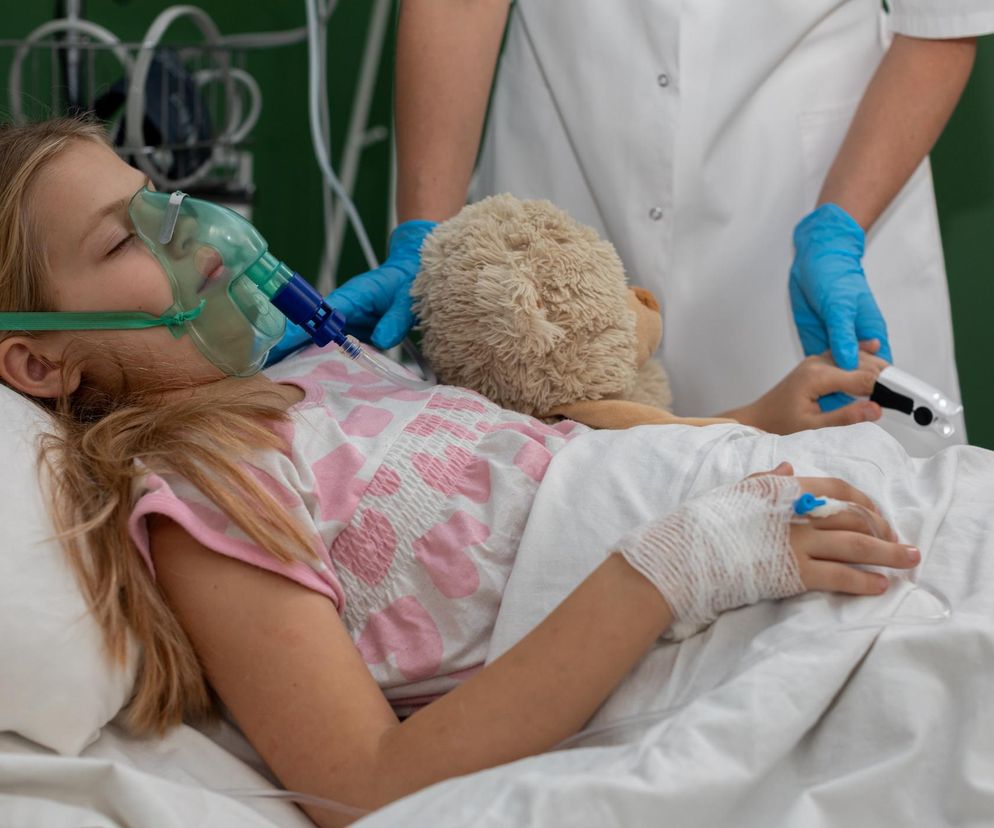Dziewczynka leżąca w szpitalnym łóżku, z maską tlenową na twarzy, obok pluszowa zabawka. Pielęgniarka w tle trzyma pulsoksymetr. Zdjęcie symbolizuje walkę dzieci z groźnym wirusem RSV, o którym można przeczytać więcej na Poradnik Zdrowie.