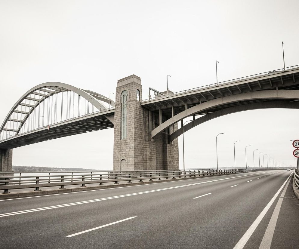 Szosa prowadzi przez długi most, który rozciąga się nad wodą w pochmurny dzień. W tle dominują dwa monumentalne łuki mostu, podparte przez solidne kamienne filary. Droga na moście ma dwa pasy ruchu, rozdzielone podwójną ciągłą linią i uzupełnione przerywanymi liniami, biegnącymi w kierunku horyzontu. Po prawej stronie szosy widoczne są dwa znaki drogowe z czerwonymi obramowaniami i liczbą 30 w środku, a także rząd latarni ulicznych o prostych, zakrzywionych słupach.