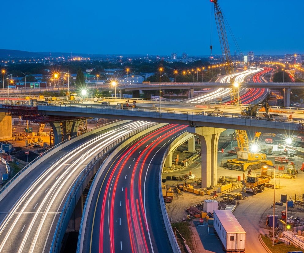 Nocny widok prezentuje rozbudowany węzeł autostradowy z wieloma pasami ruchu, po których widoczne są smugi świateł samochodów: białe od reflektorów w jednym kierunku i czerwone od świateł tylnych w przeciwnym. Nad jezdniami wznoszą się betonowe wiadukty i estakady, a w tle rozpościera się panorama miasta z licznymi światłami. Poniżej estakad trwają intensywne prace budowlane, z widocznymi maszynami budowlanymi, takimi jak koparki i dźwigi, oświetlonymi przez jaskrawe, ciepłe światła. Całość spowita jest w odcienie granatu nocnego nieba, kontrastującego z pomarańczowym blaskiem latarni ulicznych i świateł roboczych.
