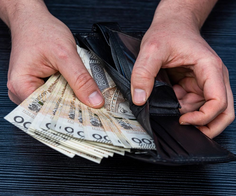Dłonie mężczyzny trzymające otwarty czarny portfel, z którego wystają banknoty o nominale 200 złotych. Zdjęcie symbolizuje kwestie finansowe związane z emeryturami, o których można przeczytać na portalu Poradnik Zdrowie.