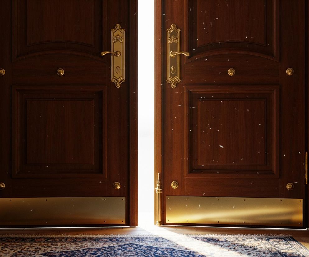 Dwoje brązowych, drewnianych drzwi otwiera się na oświetloną przestrzeń, tworząc jaskrawy, pionowy pas światła pośrodku. Drzwi ozdobione są złotymi, klasycznymi klamkami oraz złotymi listwami u dołu. Na pierwszym planie widoczny jest wzorzysty dywan w odcieniach niebieskiego i beżu, częściowo oświetlony blaskiem zza drzwi. Drobne, białe punkty unoszą się w powietrzu, sugerując kurz lub pyłki.
