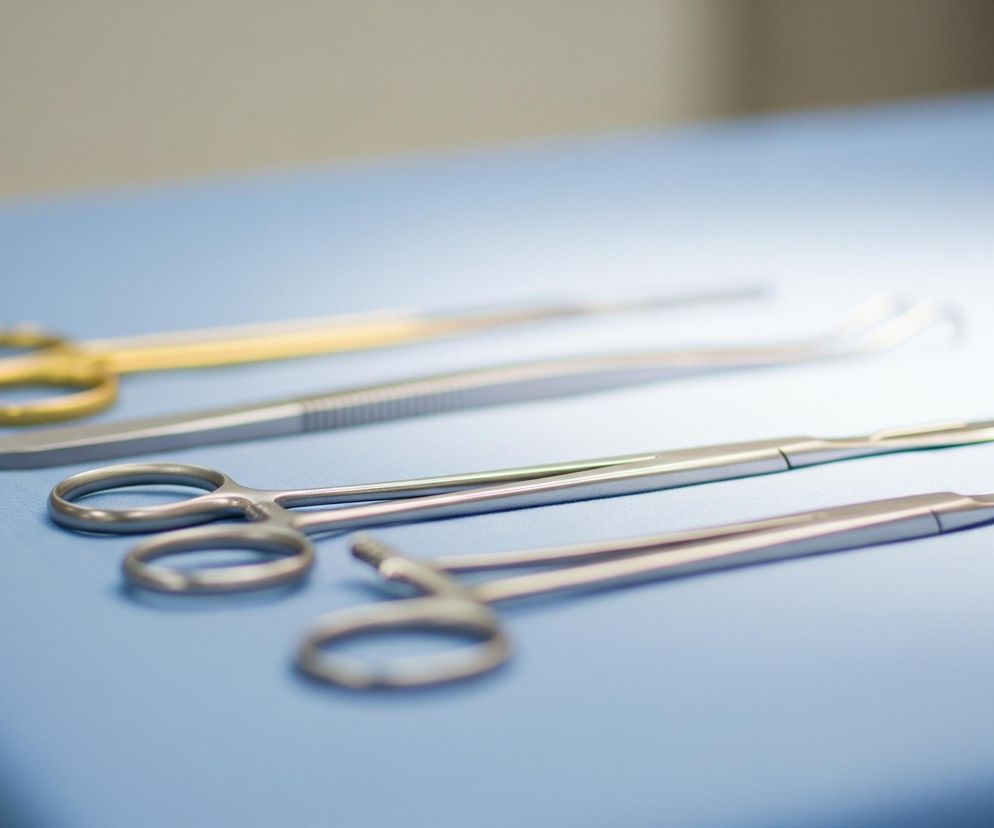 Na niebieskim, teksturowanym materiale leżą cztery metalowe instrumenty, ułożone równolegle pod lekkim kątem od lewego dolnego rogu do prawego górnego. Instrumenty to (od góry): nożyczki ze złotymi uchwytami, szczypce z karbowanymi końcówkami, długie szczypce z pierścieniowymi uchwytami oraz skalpel z ostrzem skierowanym w prawo. Tło jest rozmyte, jaśniejsze niż stół, z widocznym intensywnym źródłem światła w górnej środkowej części.