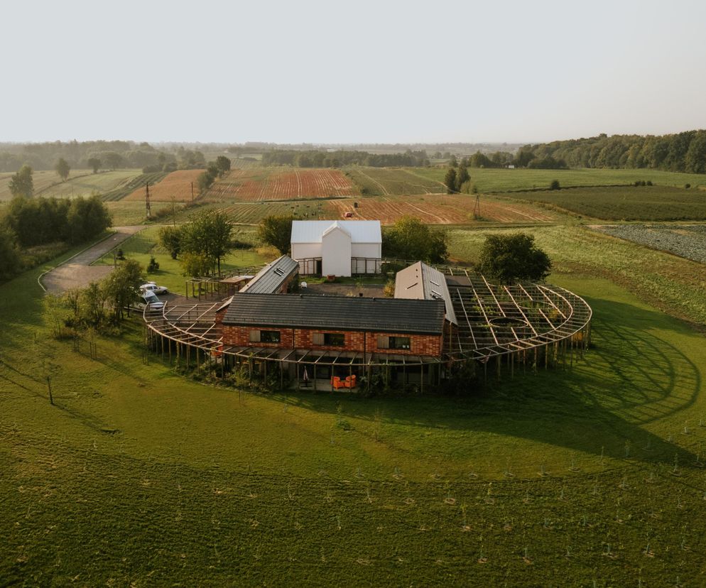Agroturystyka W Polach z lotu ptaka, przedstawiająca kompleks budynków z ceglanymi elewacjami i ciemnymi dachami, otoczony drewnianą konstrukcją okalającą prywatne ogrody. W tle widoczne są pola uprawne i lasy. Więcej o tym projekcie architektonicznym przeczytasz na portalu Architektura Murator Plus.