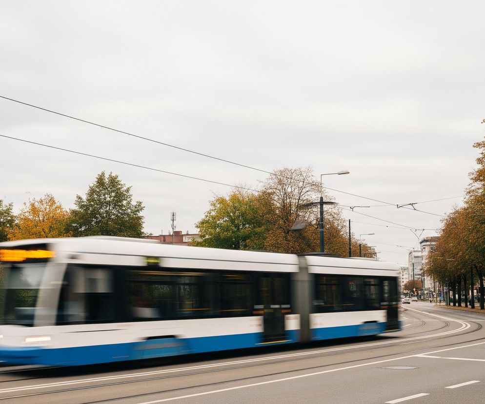 Tramwaj o białej karoserii z niebieskim paskiem u dołu pędzi po torach, pozostawiając za sobą rozmycie ruchu. Obiekt znajduje się na drodze o dwóch pasach ruchu w każdą stronę, z torami tramwajowymi pośrodku. Po obu stronach drogi rosną drzewa z jesiennymi liśćmi w odcieniach zieleni i pomarańczu, a dalej widać budynki miejskie. Niebo jest pochmurne i szare, a linie wysokiego napięcia przecinają je poziomo.