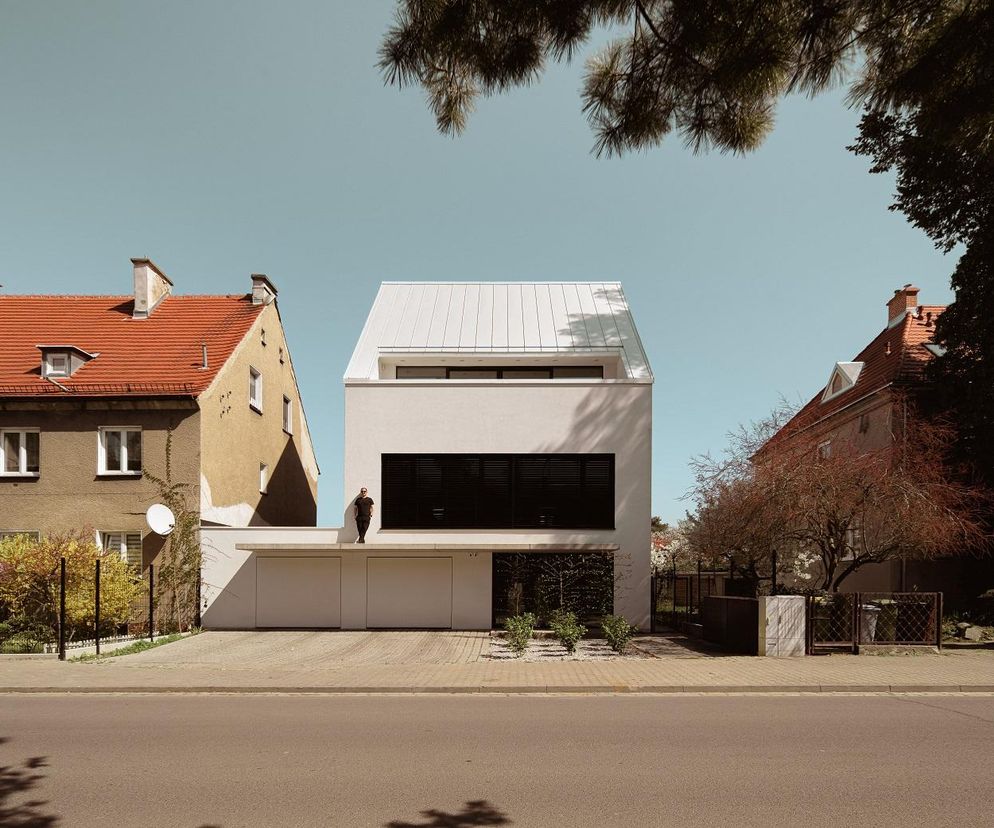 Nowoczesny biały dom jednorodzinny „dom pomiędzy” we Wrocławiu, projektu 3XA, wkomponowany między starsze budynki mieszkalne z czerwonymi dachami. Minimalistyczna fasada z czarnymi żaluzjami i metalowym dachem, widoczna na portalu Architektura Murator Plus.