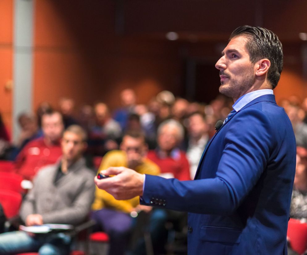 Mężczyzna w eleganckim garniturze z mikrofonem przy krawacie przemawiający do publiczności, z pilotem w dłoni, podczas spotkania biznesowego lub szkolenia. W tle widoczni słuchacze, co odzwierciedla tematykę life i biznes coachingu, o czym więcej przeczytasz na Poradnik Zdrowie.
