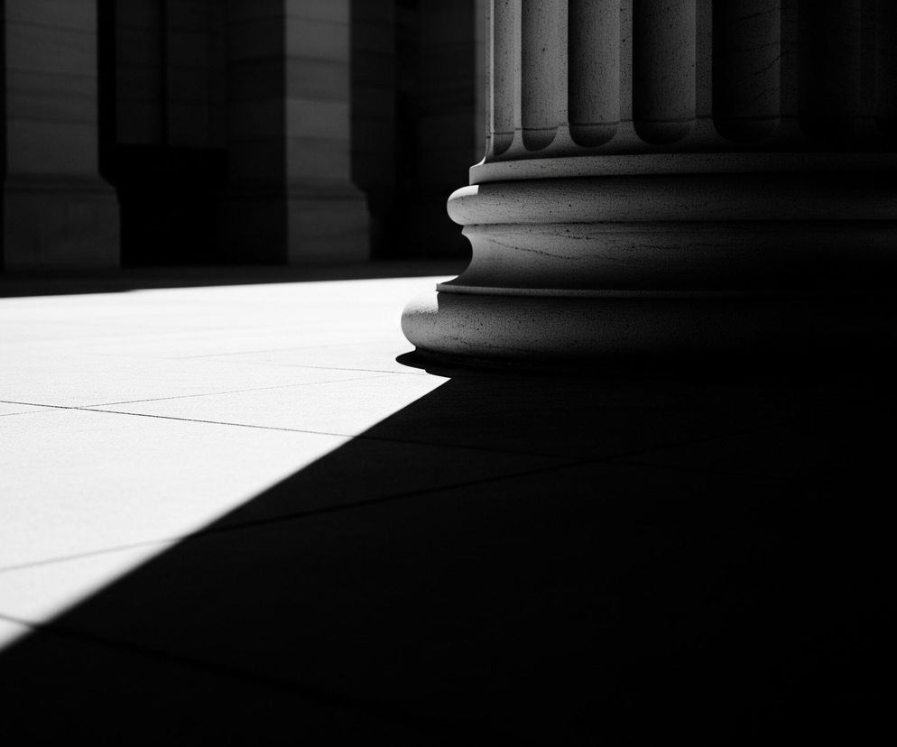 Prawa strona obrazu zdominowana jest przez podstawę masywnej, kamiennej kolumny w stylu klasycznym, która jest silnie teksturowana i żłobiona pionowo. Jasne światło pada z lewej strony, tworząc długi, ostry cień rozciągający się ukośnie przez podłogę wyłożoną jasnymi płytami, z ciemnym tłem kolumn w głębi. Całość utrzymana jest w czarno-białej tonacji, podkreślającej dramatyczny kontrast światła i cienia.