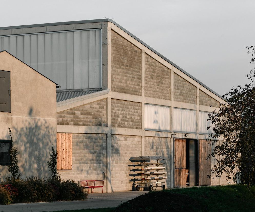 Dwa budynki gospodarcze w wiejskim krajobrazie o zachodzie słońca. Większy obiekt z widoczną betonową konstrukcją i ścianami z bloczków, z drewnianymi wrotami i świetlikami. Mniejszy budynek obok z otynkowaną elewacją. Cienie drzew i krzewów na ścianach podkreślają naturalny kontekst. Więcej o takich obiektach przeczytasz na Architektura Murator Plus.