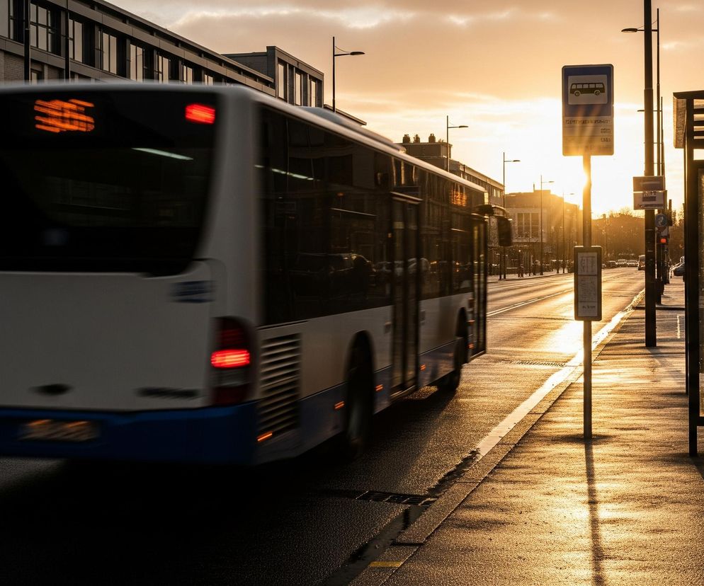 Biało-niebieski autobus z zamazanym tyłem, wskazującym na ruch, jedzie wzdłuż ulicy podczas zachodu słońca. Po prawej stronie drogi znajduje się przystanek autobusowy z wiatą i ławką, a także słup z niebieskim znakiem przystanku autobusowego, z którego bije światło zachodzącego słońca. Droga jest mokra i odbija pomarańczowo-złote światło, a w tle widać ciemne budynki i drzewa bez liści.
