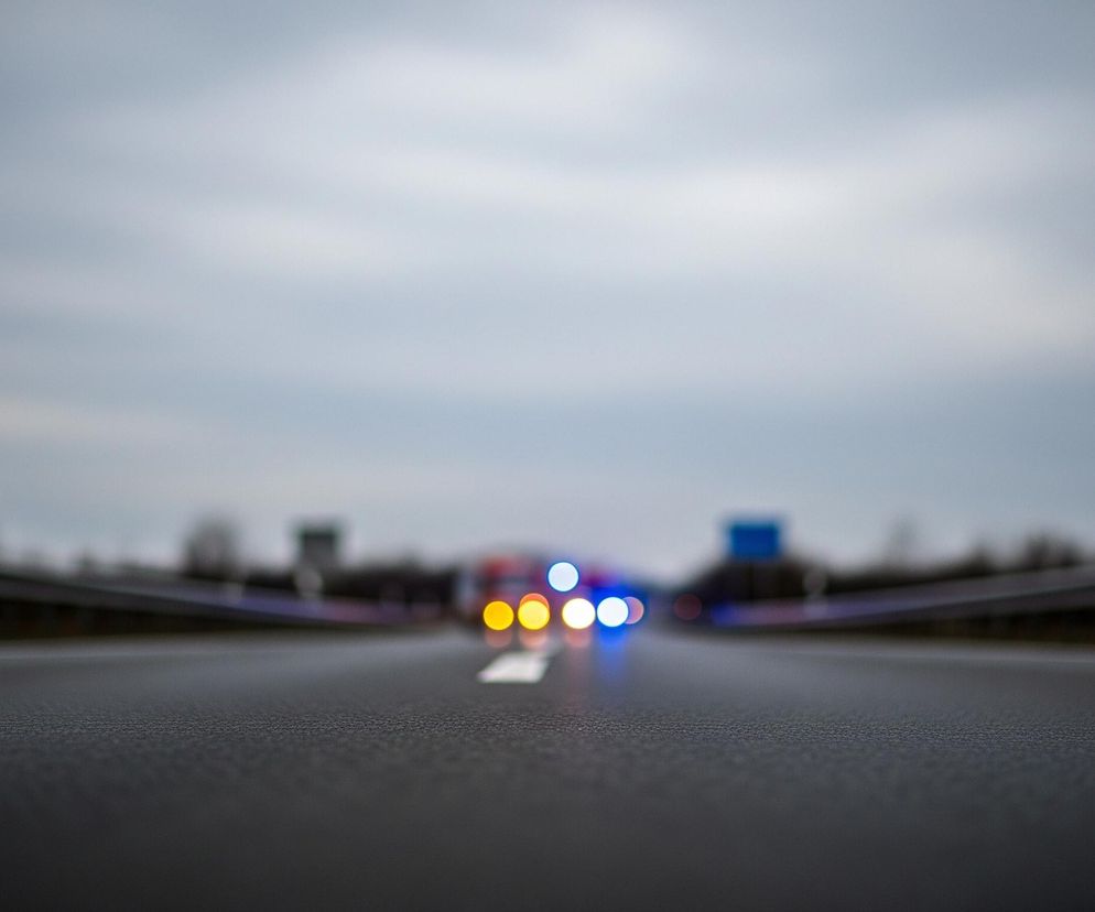 Asfaltowa nawierzchnia drogi w ciemnoszarym kolorze zajmuje dolną połowę kadru, a jej faktura jest wyraźna na pierwszym planie. Wzdłuż środka drogi, na odległym planie, widać rozmyte, jasne światła: cztery żółte po lewej stronie i trzy niebieskie po prawej, sugerujące pojazd uprzywilejowany. Droga jest otoczona metalowymi barierkami po obu stronach, które rozciągają się w głąb obrazu. Nad barierkami rozciąga się niewyraźny krajobraz z ciemnymi elementami, a horyzont zamyka jednolite, szare niebo z delikatnymi jaśniejszymi smugami.