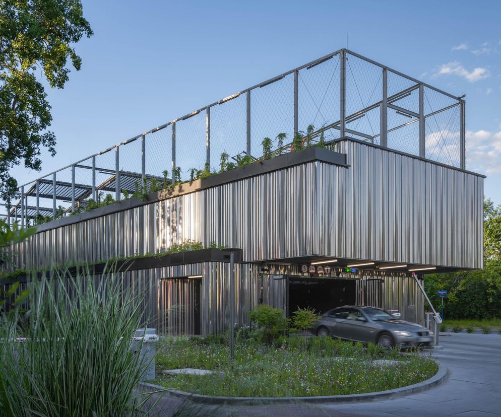 Nowoczesny garaż wielostanowiskowy projektu Medusa Group dla CD Projekt, w Warszawie. Budynek o elewacji z perforowanej blachy posiada strefę rekreacyjną na dachu z zielenią i lekkim, ażurowym ogrodzeniem. Wejście na parking z widocznym autem, otoczone roślinnością, nawiązuje do ekologicznych rozwiązań Architektura Murator Plus.