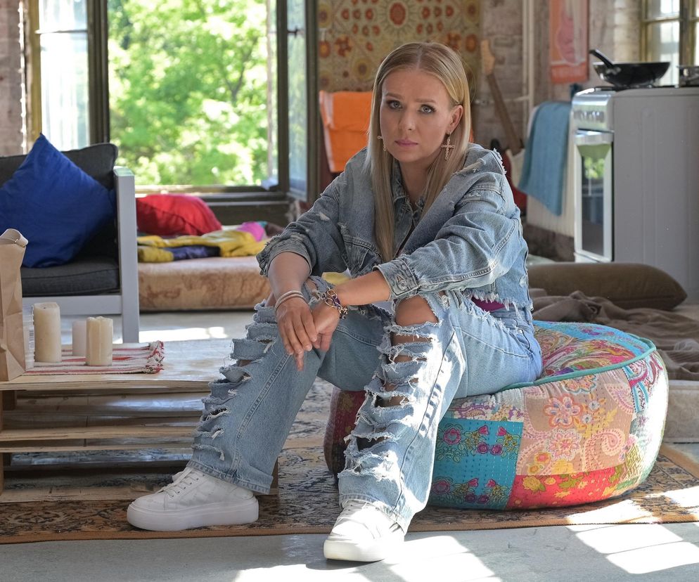 Aneta Zając jako Dominika, siedzi w zniszczonych jeansach i katanie na kolorowej pufie. Scena z serialu Pierwsza miłość ukazująca postać zamieszaną w porwanie Marysi. Więcej o wątku i innych serialach na Super Seriale.