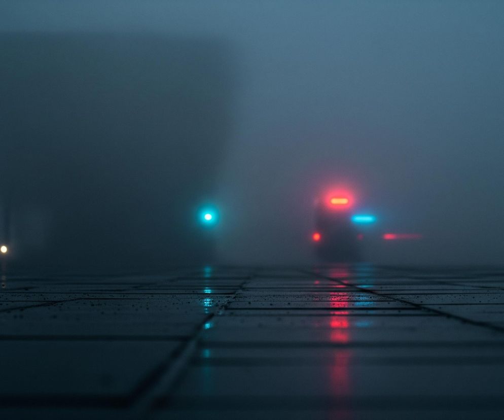 Scena przedstawia zamglone, ciemne otoczenie w odcieniach szarości i granatu. Na pierwszym planie widoczna jest mokra powierzchnia, przypominająca chodnik lub plac, pokryta kałużami i odbijająca światła. W oddali, przez gęstą mgłę, przebijają się światła: po lewej stronie jasny, turkusowy punkt świetlny, a po prawej bardziej złożony układ czerwonych i niebieskich świateł, sugerujący pojazd z włączonymi sygnałami.
