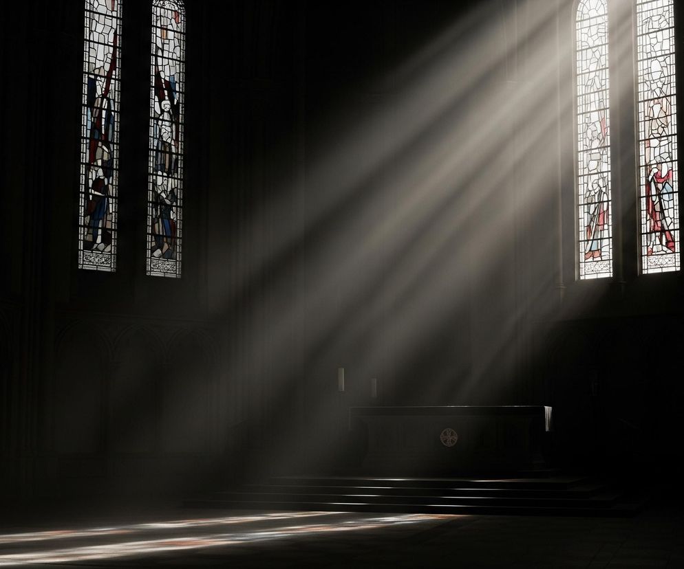 Scentralizowane, jasne promienie światła, opadające pod kątem z prawej górnej części, rozświetlają przestronne, ciemne wnętrze, prawdopodobnie sakralne. Z prawej strony widoczne są dwa wysokie, wąskie okna z kolorowymi witrażami, z których jedno jest w całości oświetlone, a drugie częściowo. Po lewej stronie widać dwa podobne okna, również z witrażami, lecz w większości zaciemnione. W centralnej części obrazu znajduje się ciemny ołtarz z ozdobnym elementem przypominającym krzyż lub ornament, a za nim dwie smukłe, jasne świece. Pod ołtarzem widać schody, a na podłodze, częściowo oświetlonej promieniami, delikatnie odbijają się barwne refleksy z witraży.