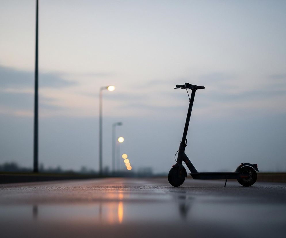 Czarna hulajnoga elektryczna stoi na mokrej nawierzchni asfaltowej drogi, która odbija światło z oddalonych latarń ulicznych, tworząc podłużne, pomarańczowe smugi. W tle widoczny jest szary, zachmurzony zmierzch. Po lewej stronie zdjęcia wznosi się ciemny słup oświetleniowy, a dalej w perspektywie, wzdłuż drogi, widać szereg zapalonych lamp, których światło jest rozmyte.