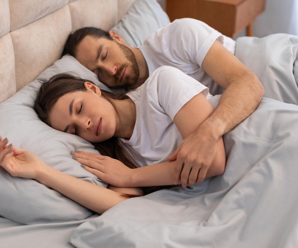 Młoda para śpi spokojnie w łóżku, przytulając się pod jasną kołdrą. Kobieta leży na brzuchu, twarzą w poduszkę, z mężczyzną obejmującym ją od tyłu. Ta scena symbolizuje regenerację i komfort, o których możesz przeczytać na portalu Poradnik Zdrowie.