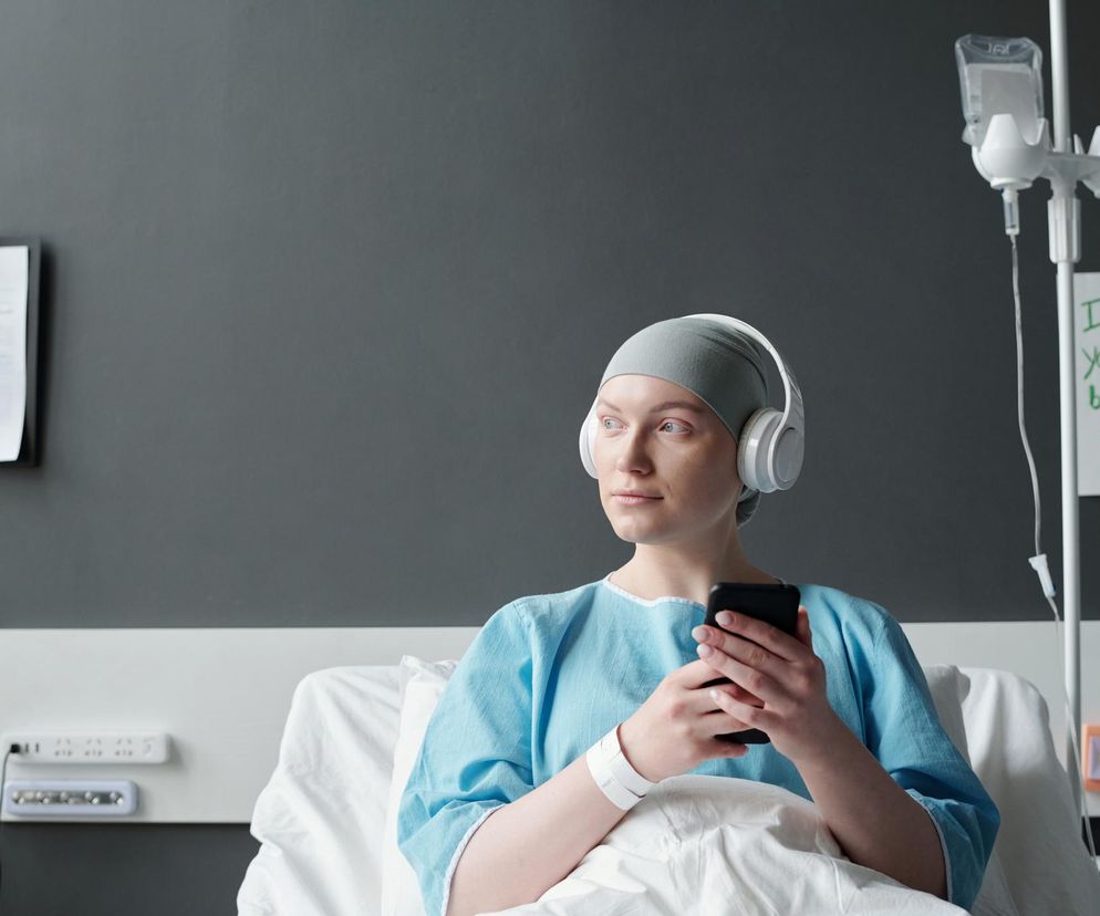 Młoda kobieta w czepku i szpitalnej koszuli leży w łóżku, trzymając smartfon i słuchawki na uszach, spogląda w prawo. Obok kroplówka i kartka z napisem „I hope you feel better soon!”. Obraz symbolizuje wyzwania zdrowotne i znaczenie profilaktyki raka u młodych dorosłych, o czym można przeczytać na portalu Poradnik Zdrowie.
