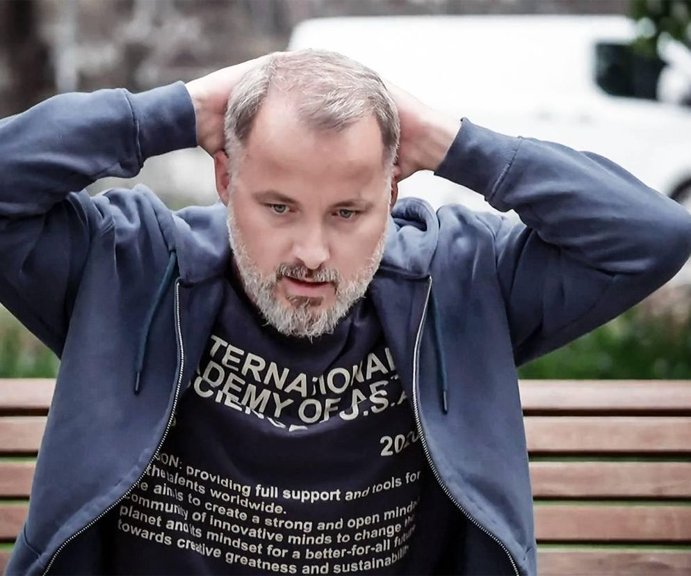 Aktor Rafał Kwietniewski jako Bartek, z dłońmi za głową, siedzi na ławce. Mężczyzna ma zatroskany wyraz twarzy, co sugeruje jego dylemat dotyczący zamieszkania z Iloną, zgodnie z treścią artykułu o serialu Pierwsza miłość, który przeczytasz na Super Seriale.