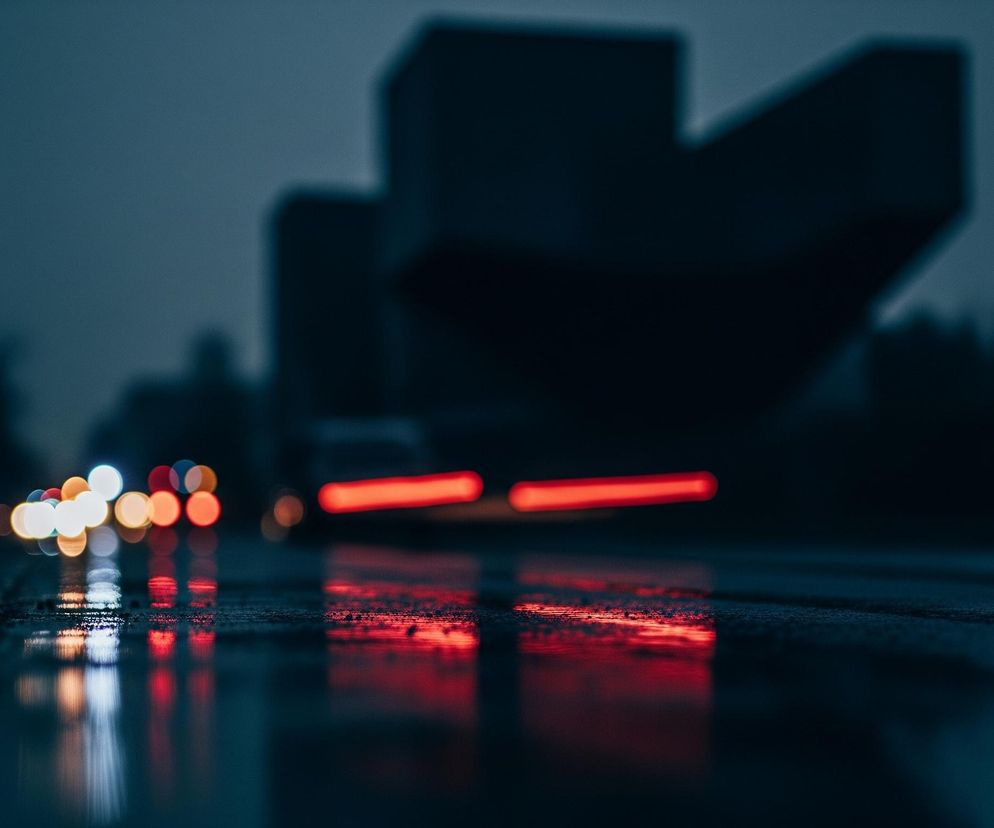 Miejski krajobraz nocą lub o zmierzchu, z mokrą asfaltową drogą na pierwszym planie, odbijającą światła. Po lewej stronie w oddali widoczne są rozmyte, okrągłe światła samochodów jadących w kierunku obserwatora, głównie w kolorach białym, żółtym i czerwonym. Po prawej stronie drogi widać dwie wyraźne, rozmyte smugi światła o intensywnym czerwonym kolorze, prawdopodobnie światła tylne przejeżdżającego pojazdu, które odbijają się w kałużach na mokrej nawierzchni. Tło stanowią ciemne, rozmyte sylwetki budynków i drzew. Całość utrzymana jest w chłodnej tonacji kolorystycznej z dominującymi odcieniami granatu i szarości.