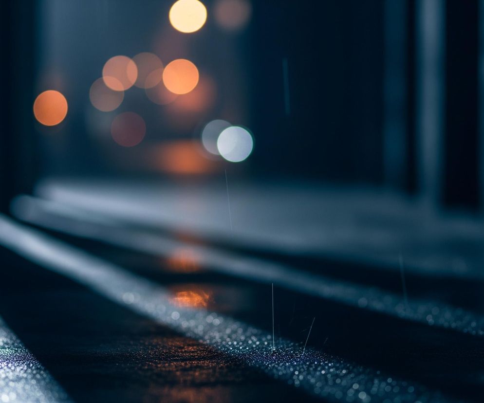 Nocna scena ulicy jest zanurzona w ciemnych, chłodnych tonach granatu i błękitu, rozjaśniona rozproszonymi, rozmyciami świateł. Deszcz pada na mokrą nawierzchnię, tworząc drobne kropelki wody, które odbijają światło, szczególnie na dwóch równoległych pasach biegnących przez środek kadru. W tle widoczne są liczne, okrągłe plamy światła o pomarańczowych i białych barwach, tworzące efekt bokeh. Pionowe smugi światła przechodzą przez lewą stronę kompozycji, natomiast po prawej stronie majaczą niewyraźne, pionowe kształty.
