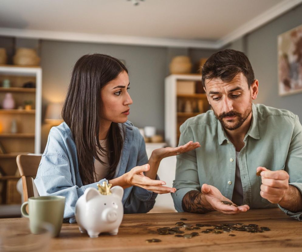 Zmartwiona para liczy monety na drewnianym stole w jasnym pokoju, obok stoi skarbonka w kształcie świnki, symbolizująca trudności finansowe Polaków, o których przeczytasz na Super Biznes. Kobieta gestykuluje, mężczyzna liczy drobne, podkreślając problem oszczędzania.
