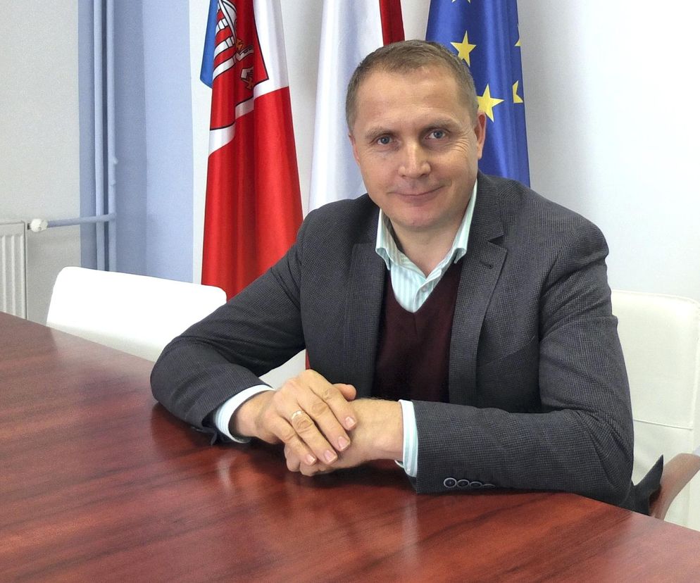 Piotr Wielgomas, wiceprezes Dawtony, siedzi przy stole, ubrany w marynarkę, sweter i koszulę. W tle widoczne są flagi Polski i Unii Europejskiej, wskazujące na kontekst polityczno-biznesowy, o którym można przeczytać na Super Biznes.