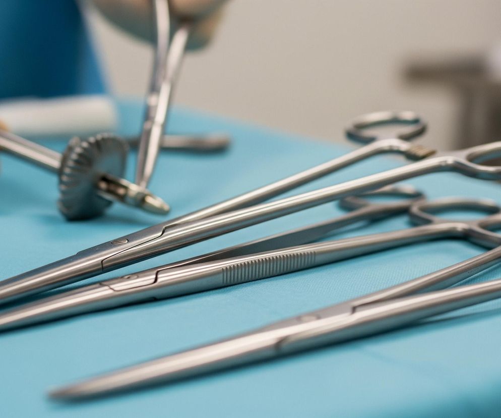 Na turkusowym blacie leży kilka metalowych narzędzi medycznych. W centrum kadru widoczne są trzy podłużne narzędzia przypominające kleszcze lub nożyczki, ułożone równolegle do siebie, z rękojeściami w kształcie owalnych pętli skierowanymi w prawą stronę. Na dalszym planie, w lewej górnej części, widać dłoń w beżowej rękawiczce chirurgicznej, trzymającą inne metalowe narzędzie z ząbkowanym okrągłym elementem. W tle, częściowo rozmyte, majaczy fragment osoby w niebieskim ubraniu oraz jasna, niezidentyfikowana przestrzeń.