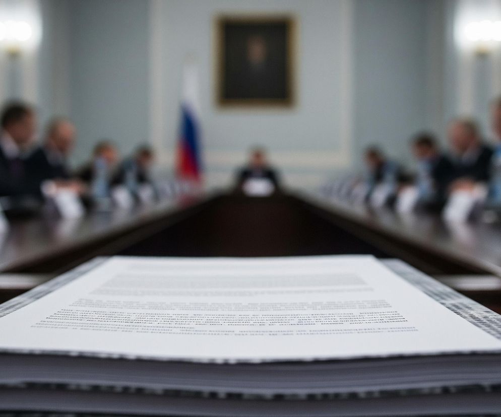 Duży, biały stos dokumentów na brązowym stole konferencyjnym, z rozmazanymi postaciami polityków na dalszym planie. Flaga Rosji i portret wiszą w tle. Dowiedz się więcej o przewidywaniach Jackowskiego na Super Biznes.