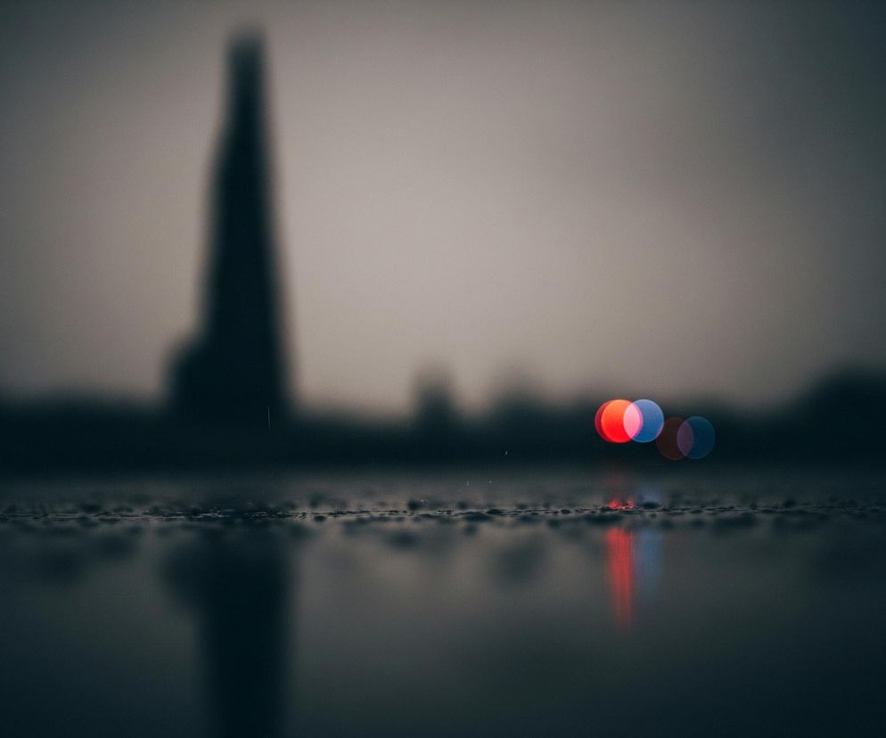 Powierzchnia pokryta kroplami wody, z wyraźnymi odbiciami światła, zajmuje dolną połowę obrazu. Powyżej niej, w tle, widoczne są cztery rozmyte punkty świetlne: dwa czerwone i dwa niebieskie, zlewające się ze sobą. W tle, jeszcze bardziej rozmyte, majaczą ciemne, pionowe kształty budynków lub innych konstrukcji. Cała scena utrzymana jest w ciemnych, stonowanych odcieniach szarości i granatu, z wyjątkiem kolorowych świateł.