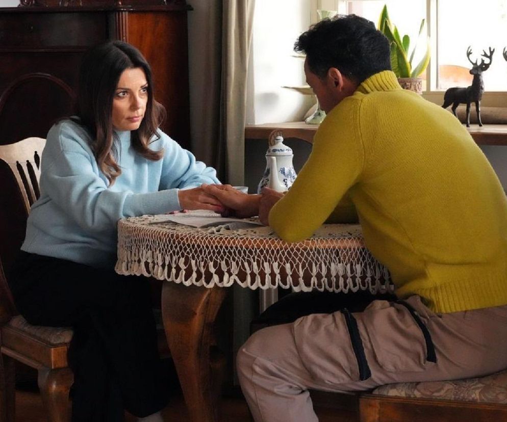 Katarzyna Glinka (Kasia) i Rafał Cieszyński (Mariusz) siedzą przy stole, trzymając się za ręce, w emocjonalnej scenie z serialu „Barwy szczęścia, której szczegóły można przeczytać na Super Seriale. W tle widać drewniane meble i rośliny doniczkowe.